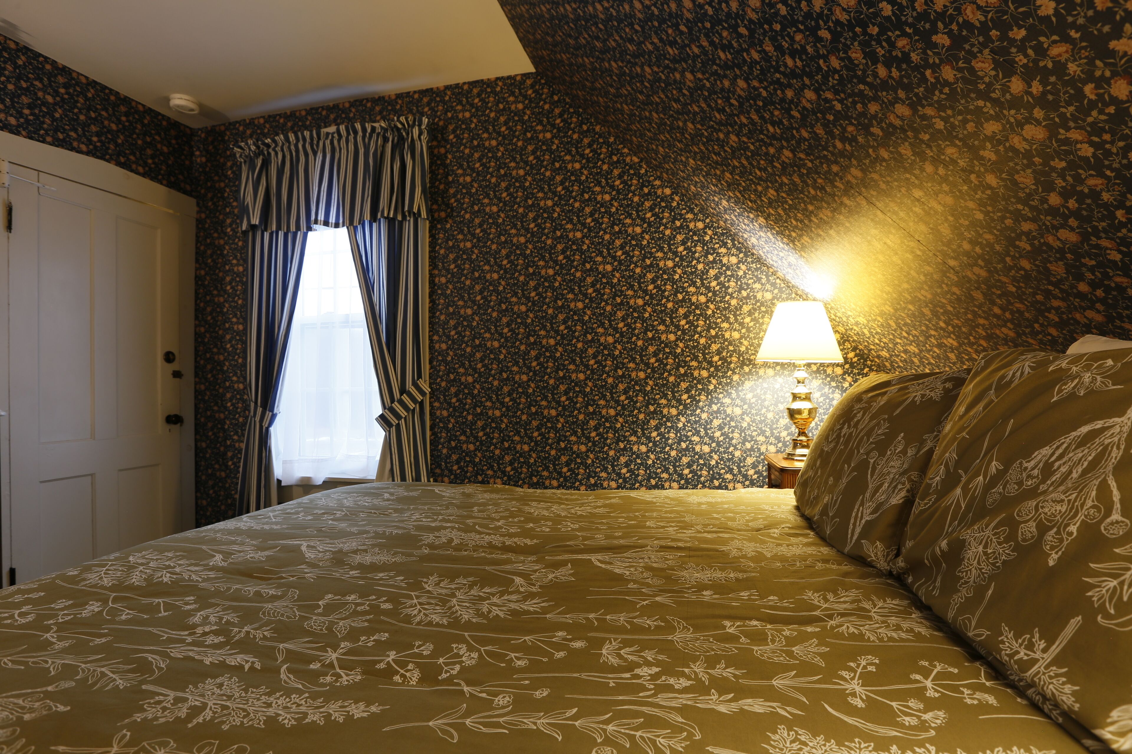 A bedroom with a floral-patterned bedspread, a window with curtains, and a lamp on a bedside table.