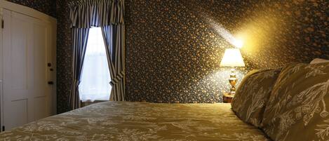 A bedroom with a floral-patterned bedspread, a window with curtains, and a lamp on a bedside table.