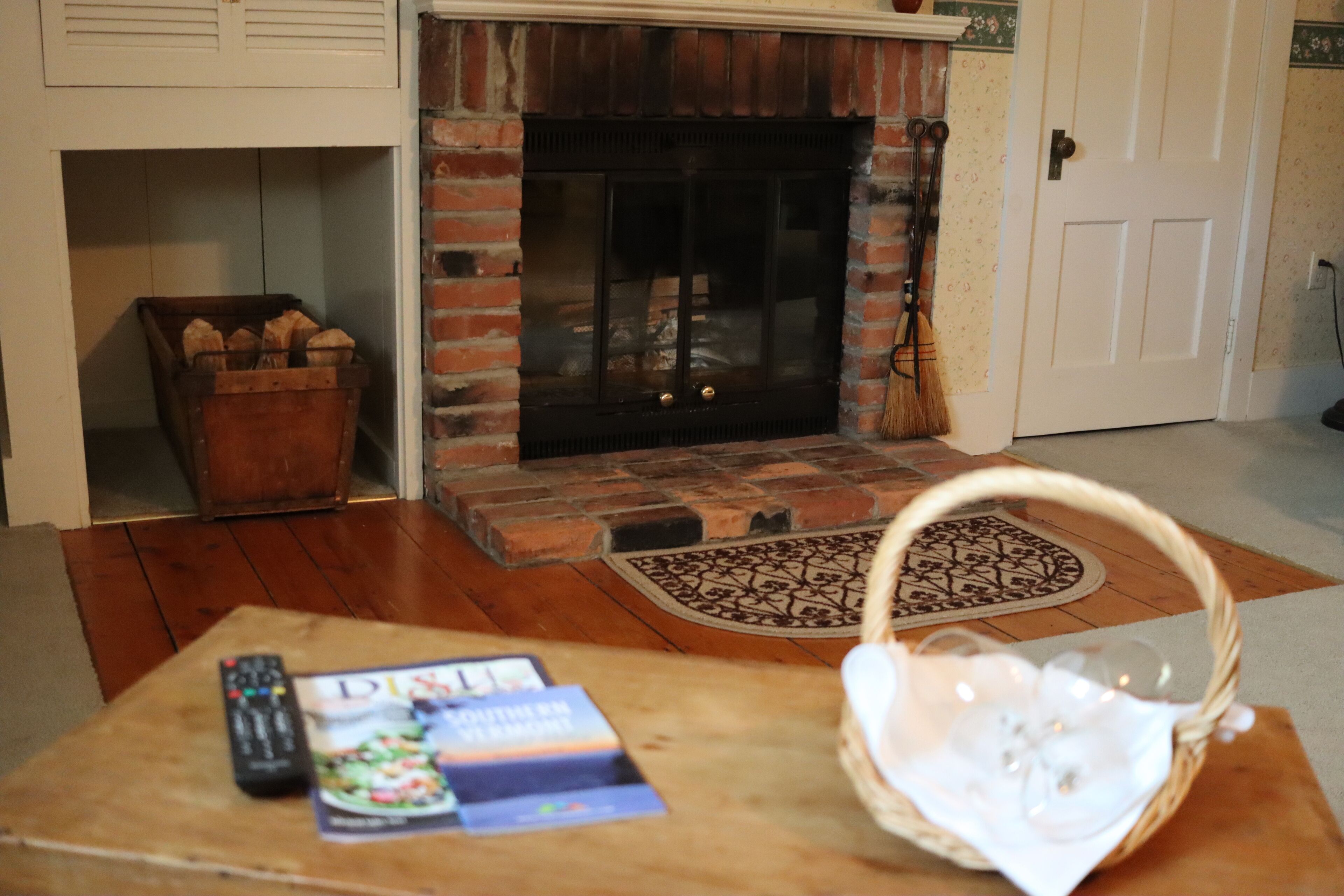 Ein Wohnzimmer mit Kamin aus Ziegelsteinen, einem Couchtisch aus Holz mit Fernbedienung und Zeitschriften sowie einem Korbwägelchen.