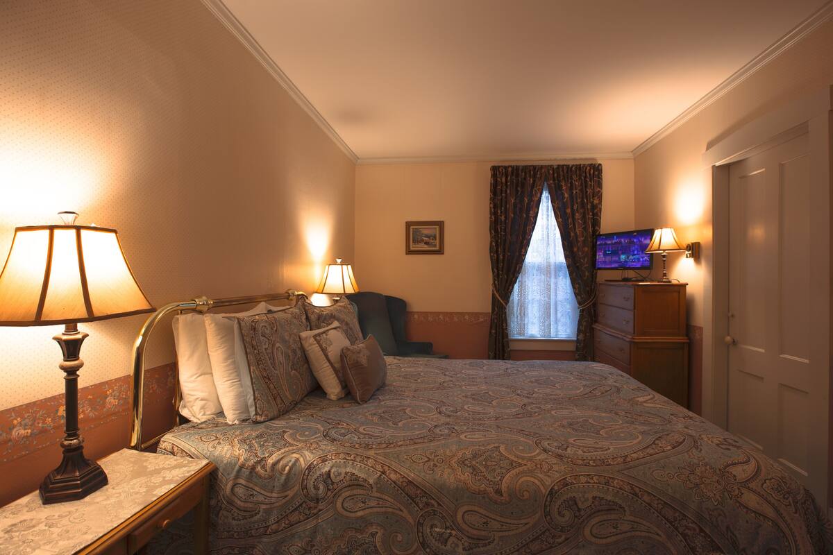 A hotel room with a bed, two bedside lamps, a television, a dresser, and a window with curtains.