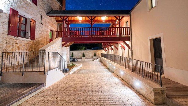 A courtyard with a wooden staircase leading to a balcony, surrounded by stone buildings with red shutters.