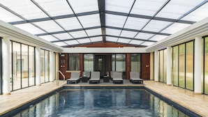 A modern indoor swimming pool with glass walls, wooden flooring, and lounge chairs.