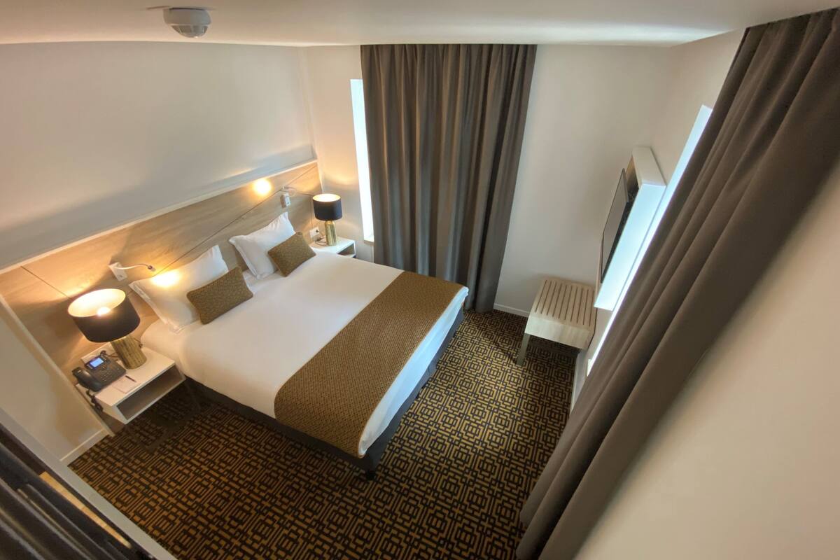 Een hotelkamer met een groot bed, nachtlampjes, een bureau met telefoon en een raam met gordijnen.