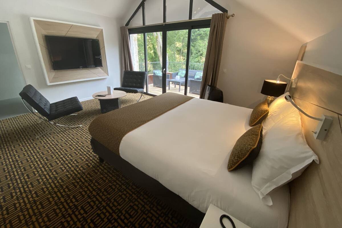 Hotelkamer met een groot bed, een flatscreen televisie aan de muur, een zithoek met een stoel en tafel, en een balkon met tuinmeubilair.