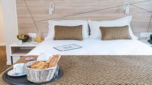 A neatly made bed with a headboard, a bedside table with a fruit bowl, a tray with breakfast items, and a basket of bread.
