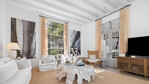 A modern living room with white furniture, a large window with curtains, a television, and a wooden cabinet.