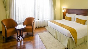 A hotel room with a large bed, two armchairs, a small table, and a window with sheer curtains. - Travellers Safari - Waridi (Nairobi)