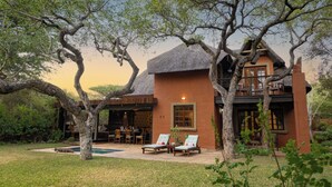 A rustic building with a thatched roof, surrounded by trees and a pool area with lounge chairs. - Braai Safaris Lodge (Hoedspruit)