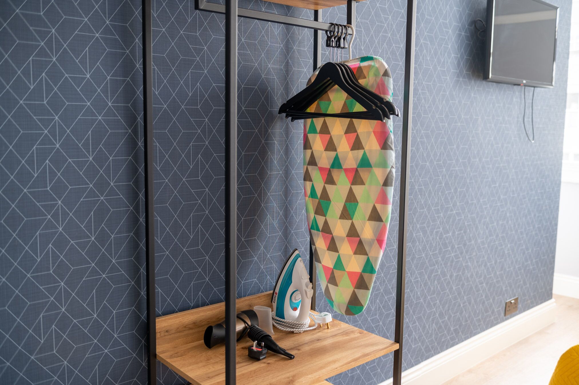 A neatly organized room with a geometric-patterned wall, a wooden shelf with an iron, a colorful garment, and a television mounted on the wall.