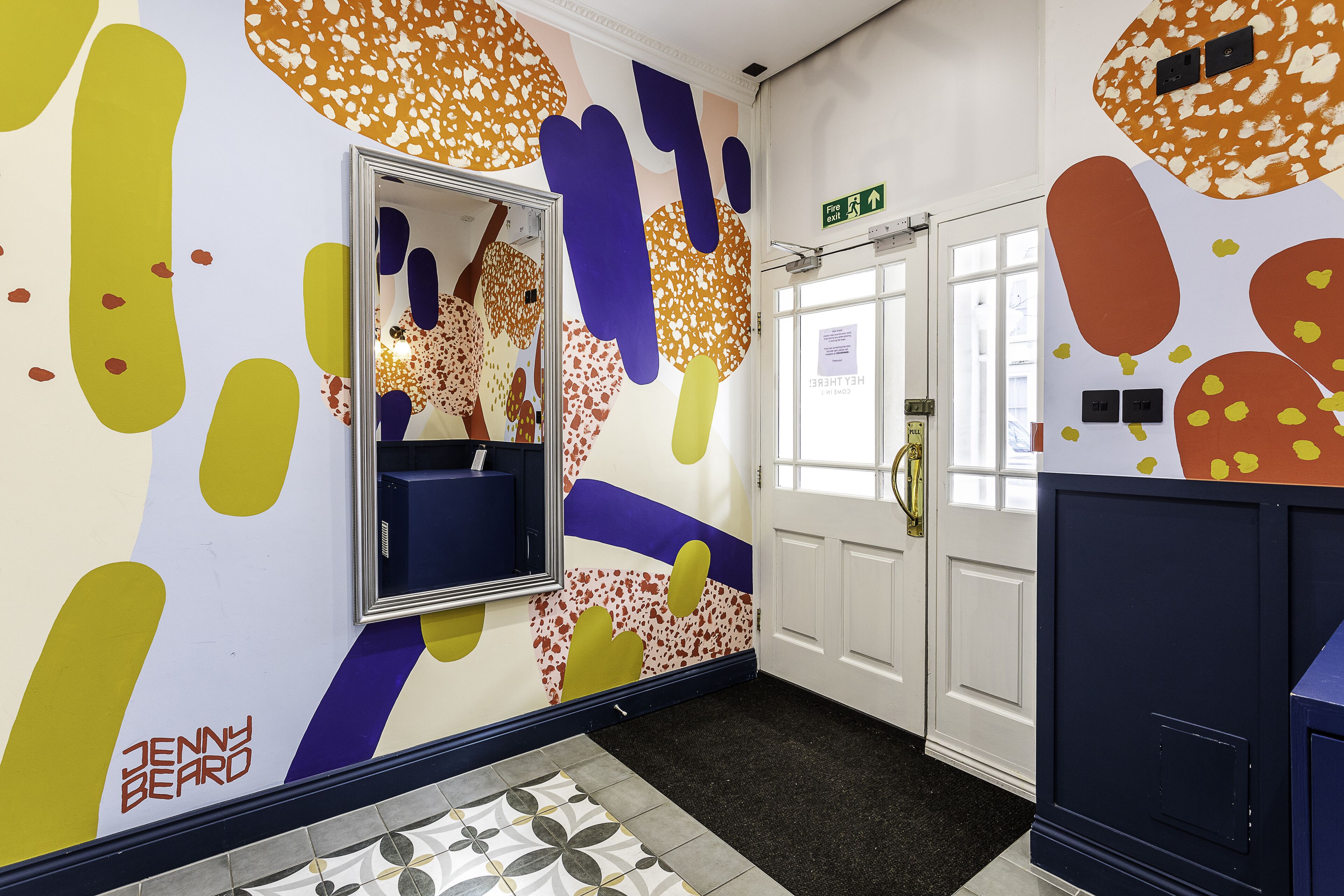 A hallway with a mirror, a door with an exit sign, and a patterned floor.