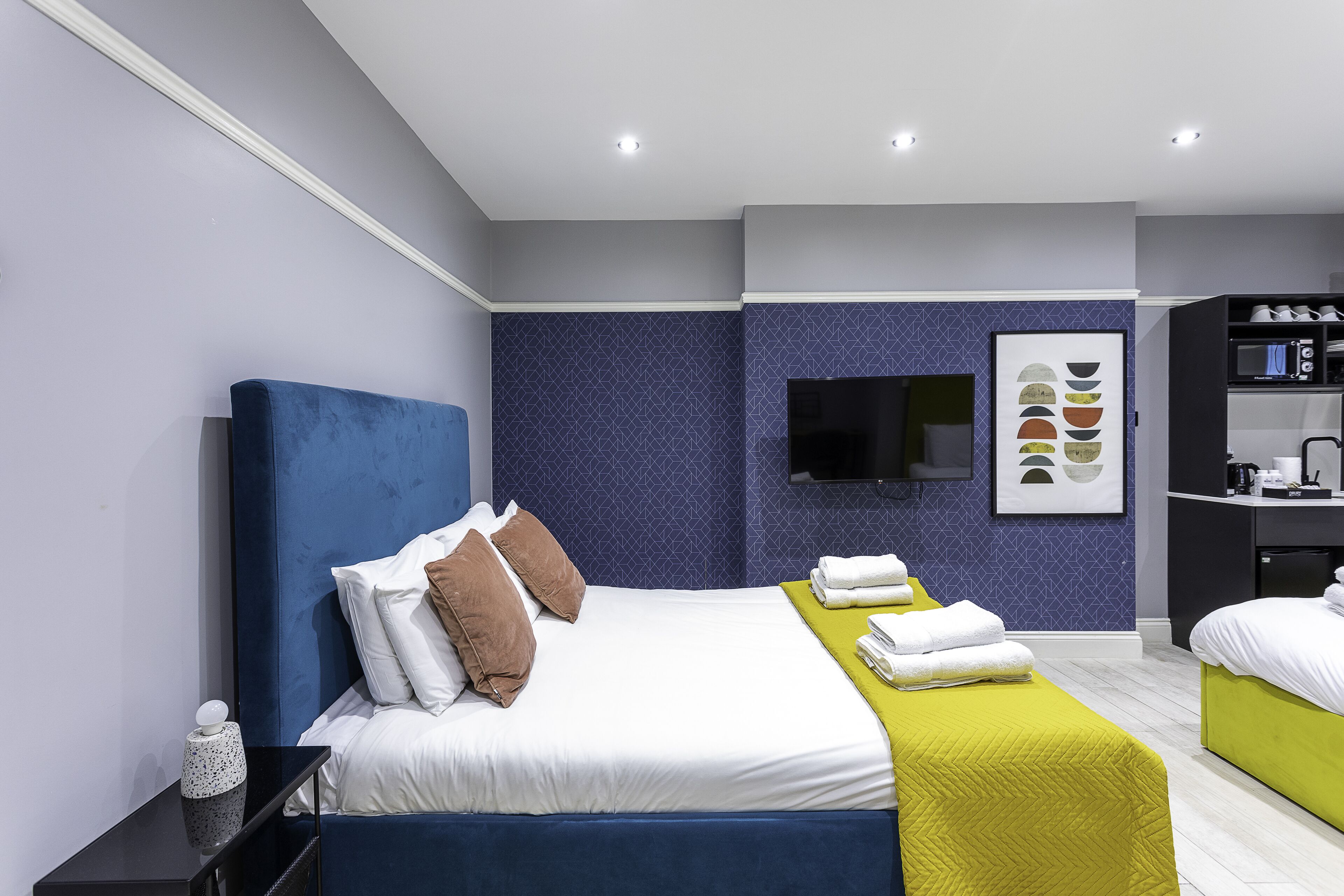 A modern hotel room with a large bed, a TV mounted on the wall, and a small kitchenette.