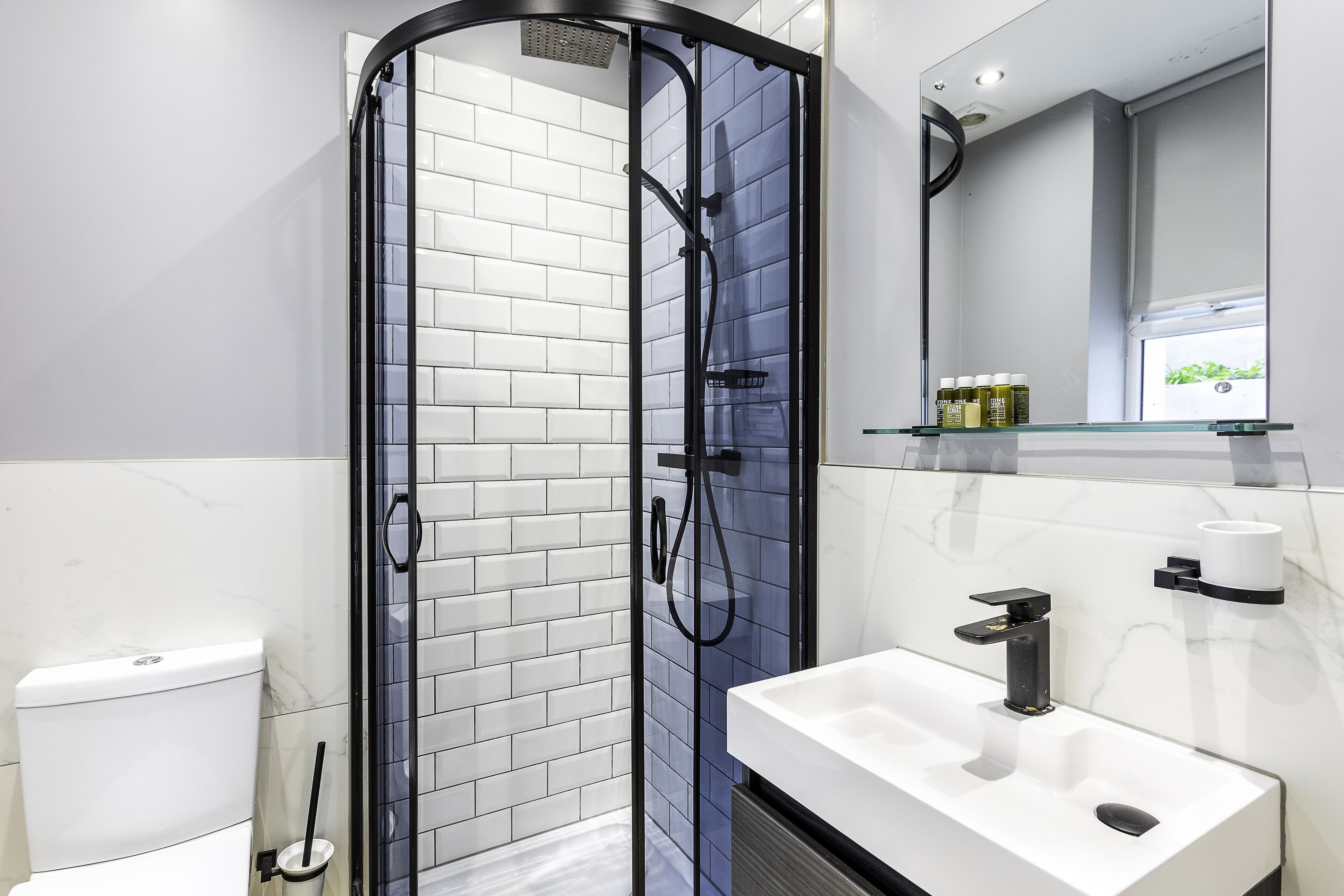 Un baño moderno con ducha acristalada, paredes cubiertas de azulejos blancos, un lavabo blanco y un espejo con un estante superior.
