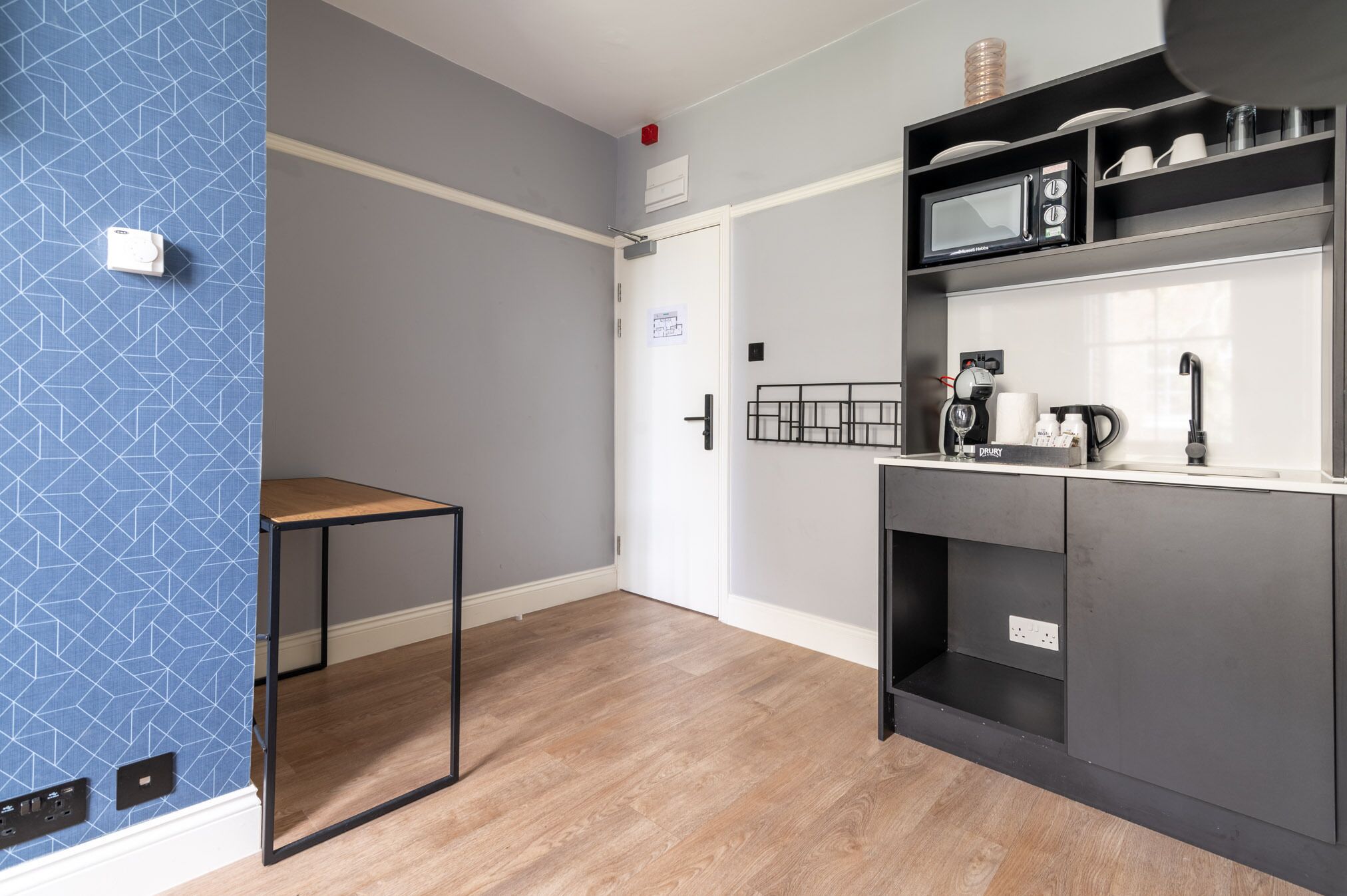Une cuisine moderne équipée d’un four à micro-ondes, d’une machine à café et d’une bouilloire sur le plan de travail. On y trouve une petite table et une porte avec une pancarte.