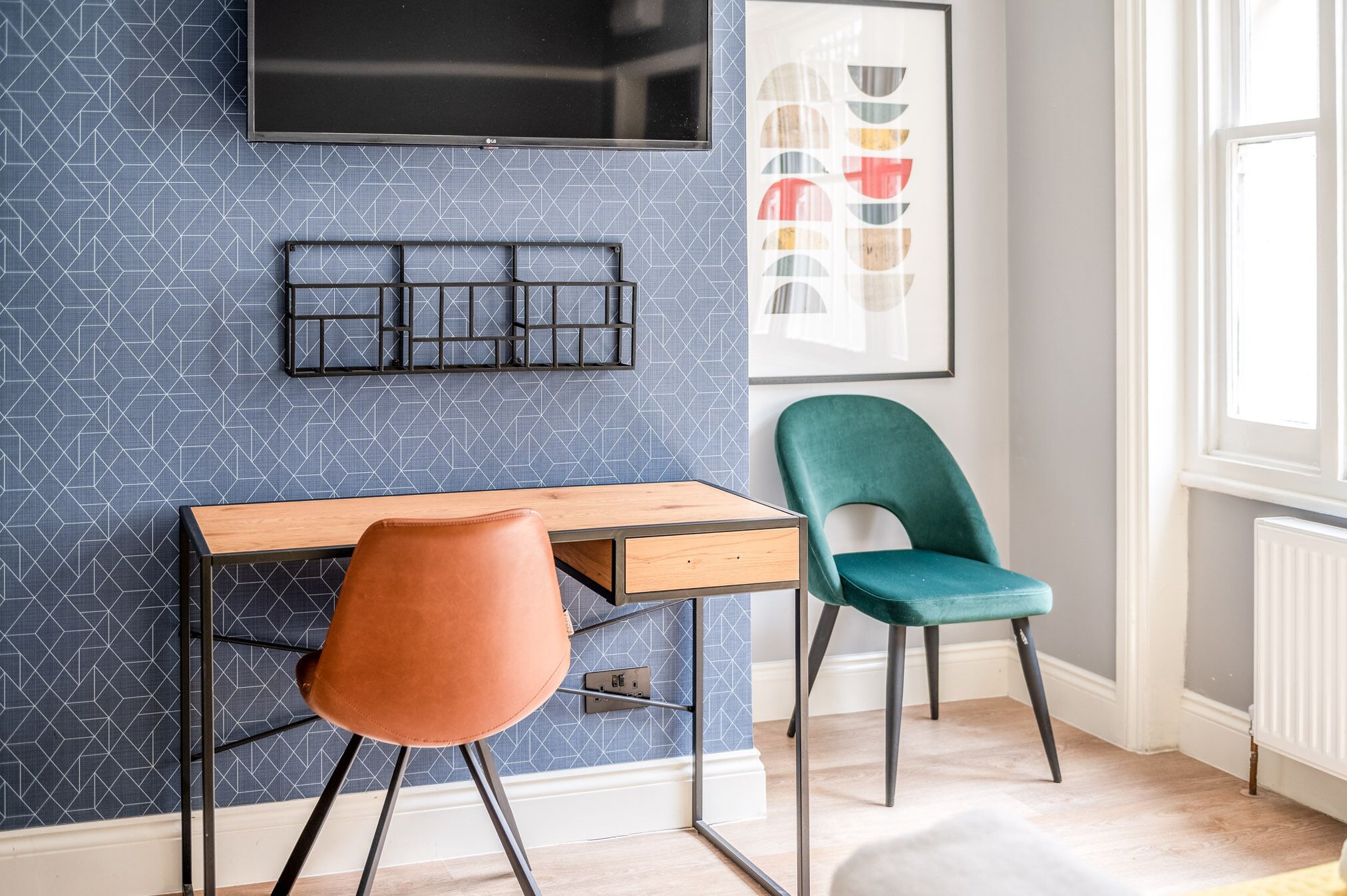 Una habitación moderna con un escritorio, dos sillas, un televisor montado y una obra de arte abstracta enmarcada.