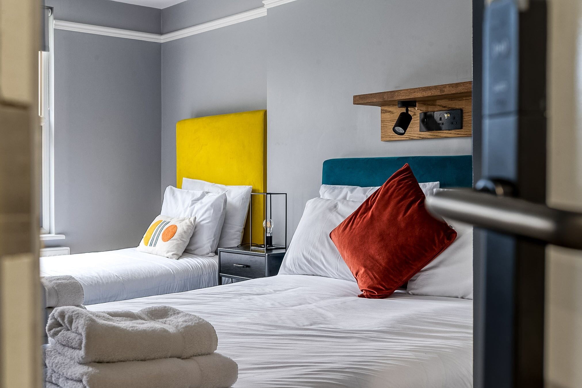 A hotel room with two beds, a wooden shelf with a lamp, and a bedside table.