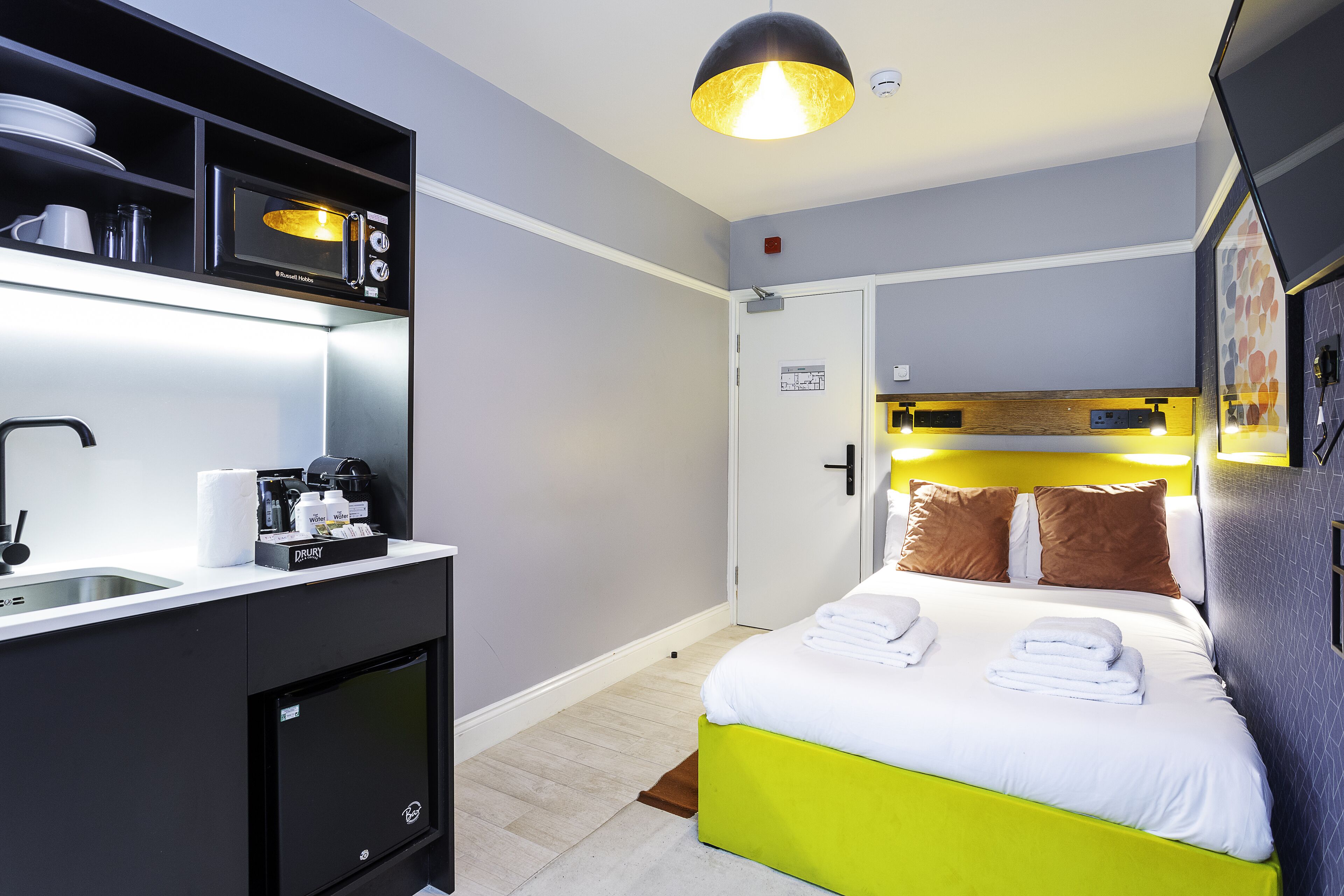 Habitación de hotel compacta con kitchenette, cama con ropa de cama blanca y verde, y una lámpara colgante moderna.