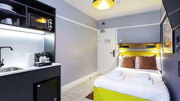 Habitación de hotel compacta con kitchenette, cama con ropa de cama blanca y verde, y una lámpara colgante moderna.