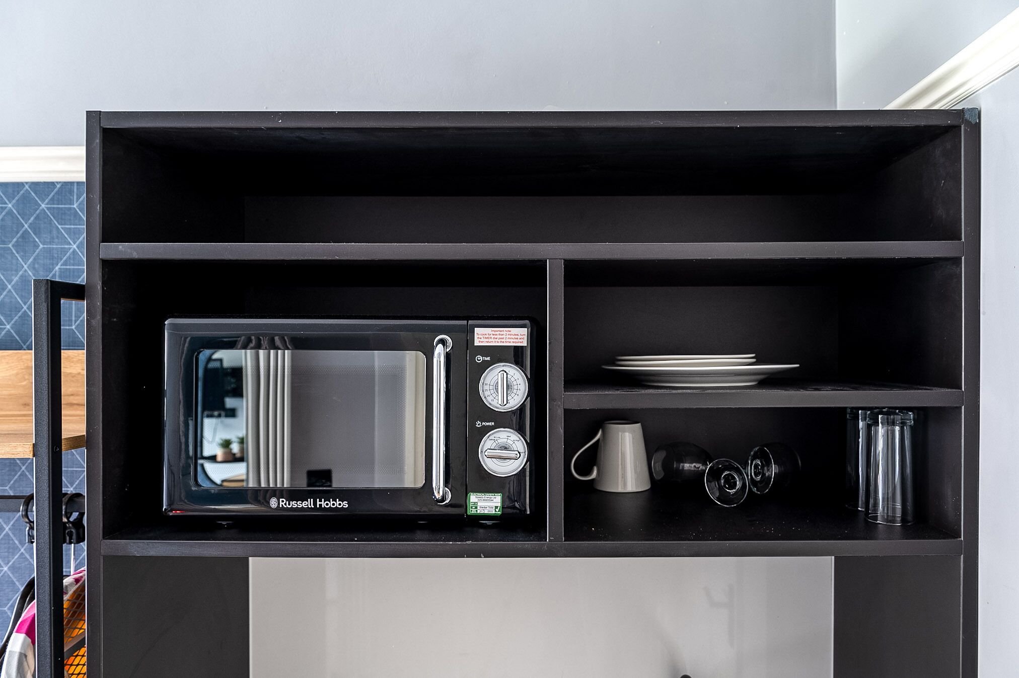 Eine kompakte Küche mit integriertem Mikrowellenherd, einem Regal mit Tellern und einer Tasse sowie einem kleinen Schrank.