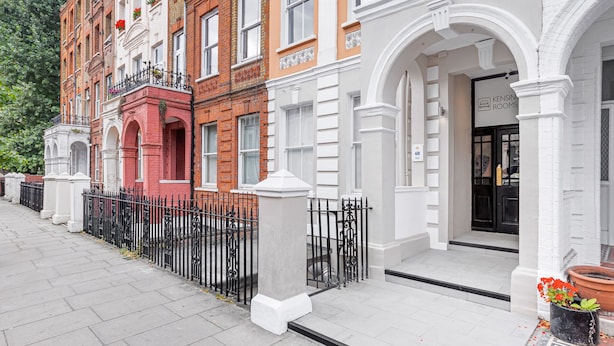 Una hilera de edificios de varios pisos con una combinación de fachadas de ladrillo y blancas, una valla de metal negro y un letrero que dice 'Kensington Rooms'.
