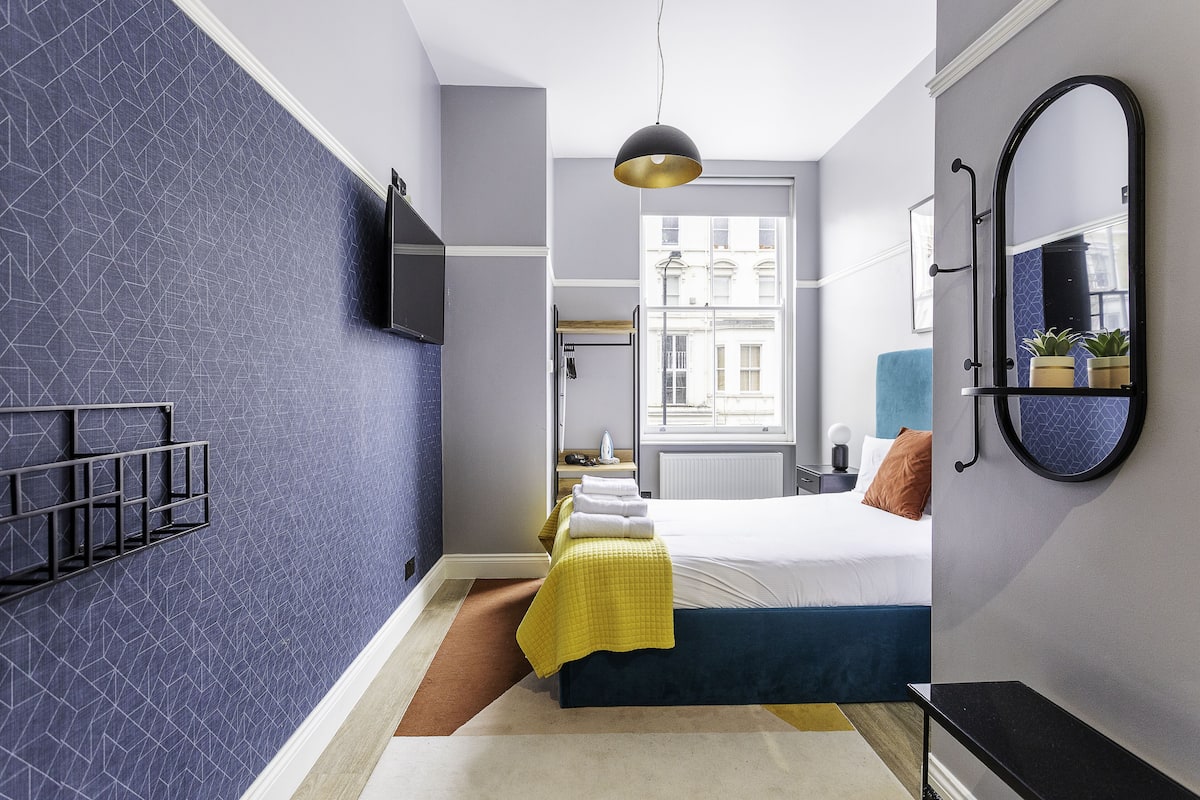 A modern hotel room with a geometric-patterned wall, a bed with a yellow and blue bedspread, a black bedside table, a round mirror, and a hanging pendant light.