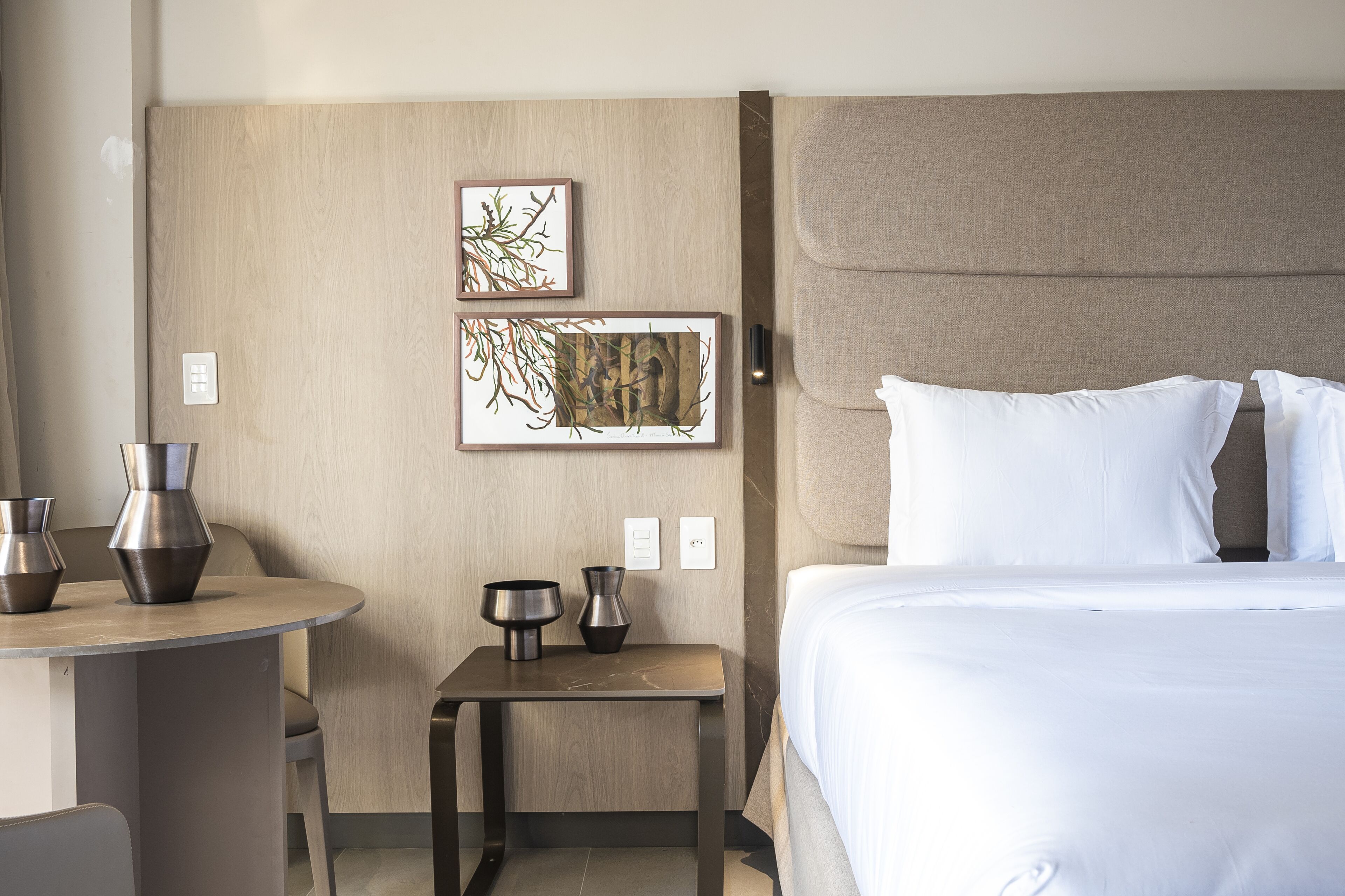 Una habitación de hotel con una cama, una mesita de noche con dos jarrones y dos obras de arte enmarcadas en la pared.