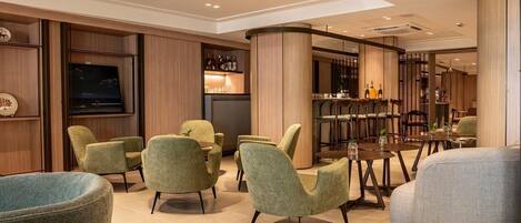 A modern lounge area with green upholstered chairs, a bar with bottles, and a television on the wall.