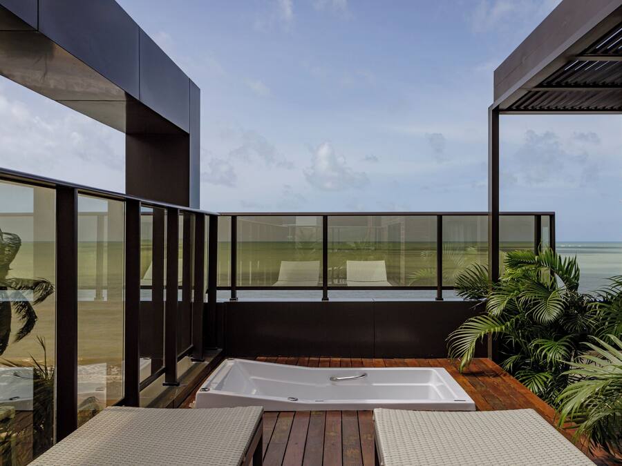 A modern outdoor bathroom with a large bathtub, wooden deck, and glass walls offering a view of the ocean.