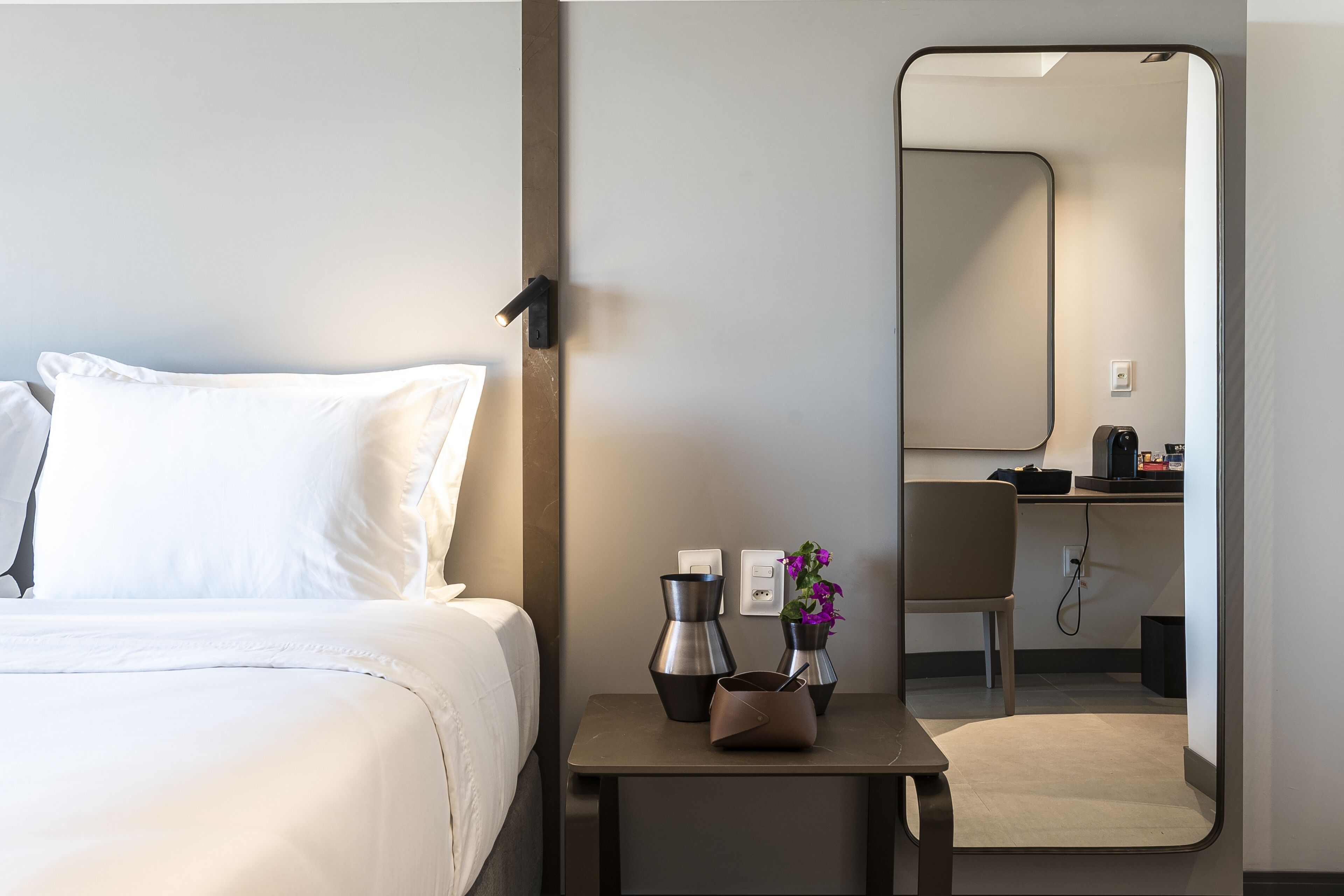 Een hotelkamer met een bed, een nachtkastje met koffiezetapparaat en een vaas met bloemen, een spiegel en uitzicht op de badkamer.