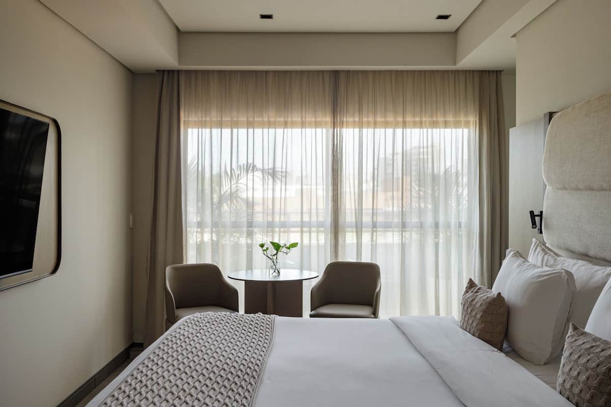 Habitación de hotel con una cama grande, dos sillones, una mesita con una planta y un ventanal con cortinas.
