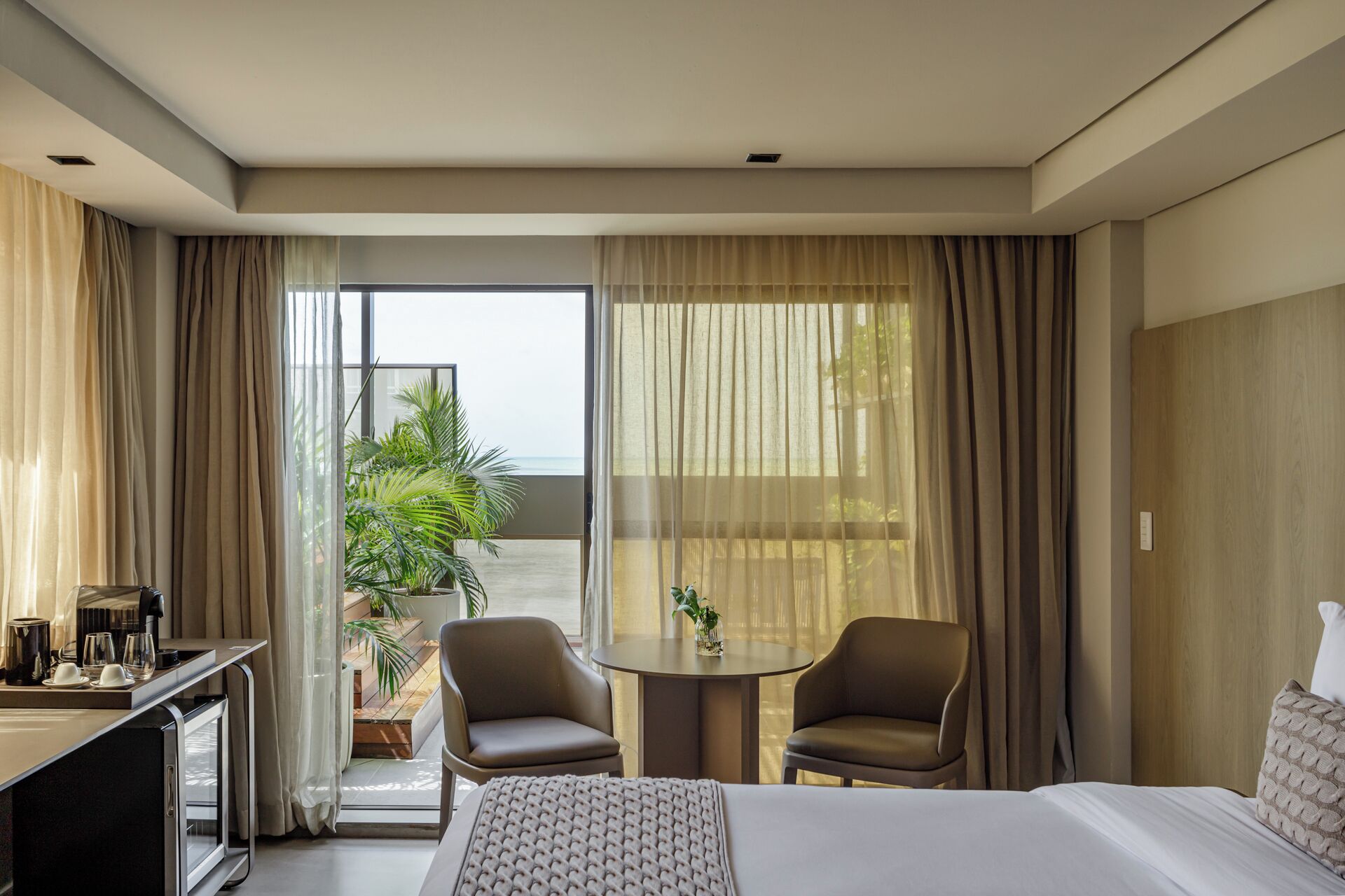 Habitación de hotel con cama, dos sillas, una mesita y un escritorio con cafetera. Cuenta con un balcón con cortinas y vistas al océano.