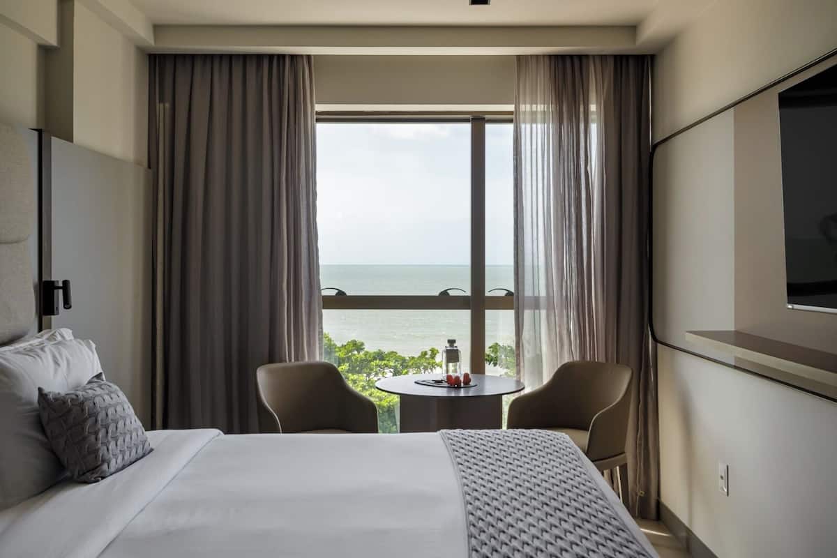 Habitación de hotel con cama, dos sillas, una mesita, televisor y ventana grande con vista al mar.