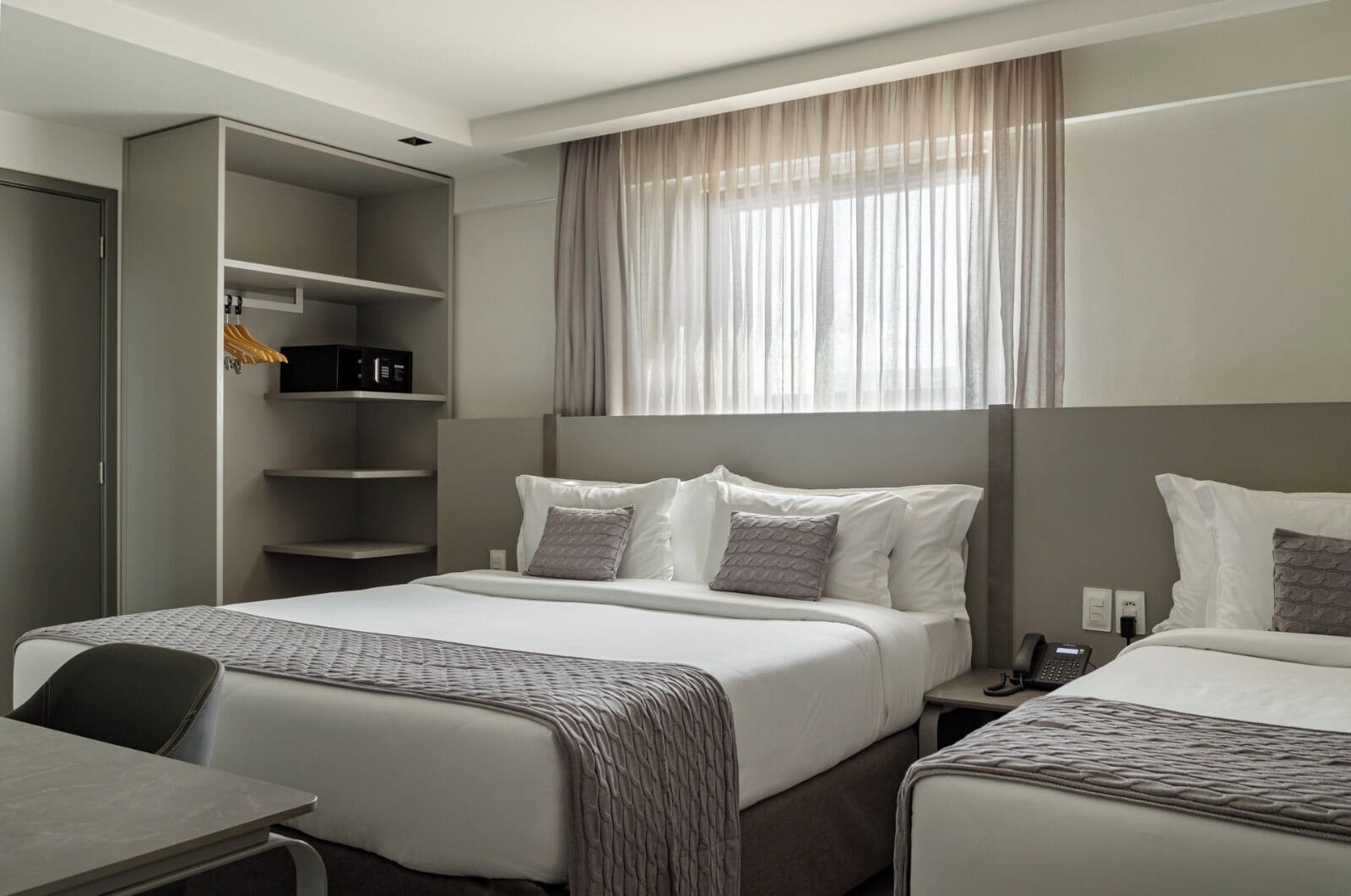 A hotel room with two beds, a desk, a wardrobe, and a large window with sheer curtains.