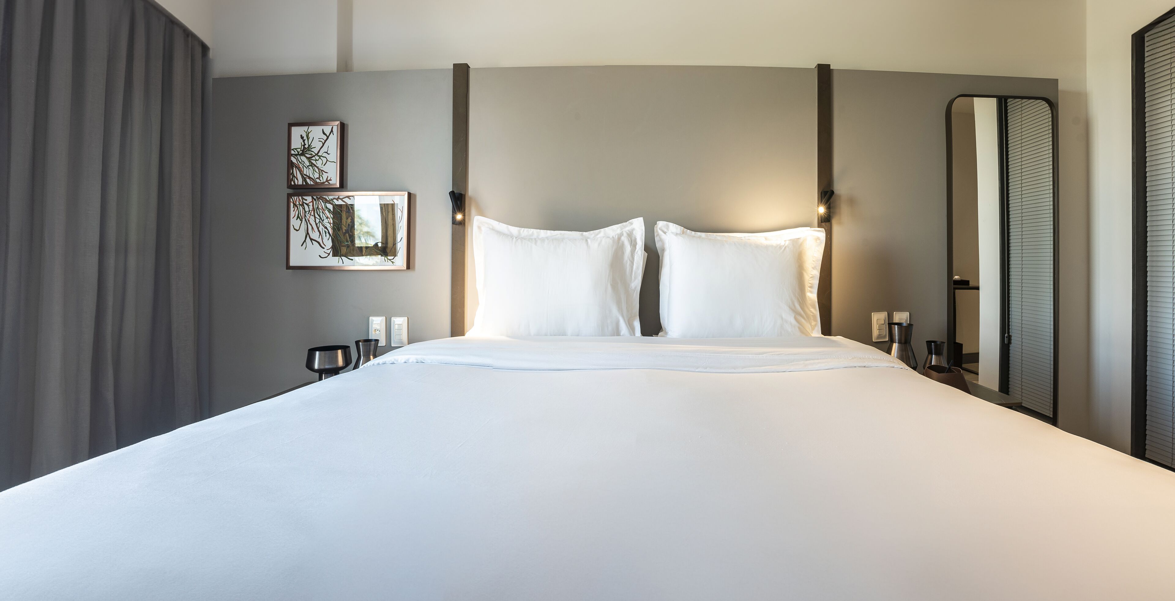 A modern hotel room with a large bed, two bedside tables, a nightstand, a wall-mounted light, and a large mirror.