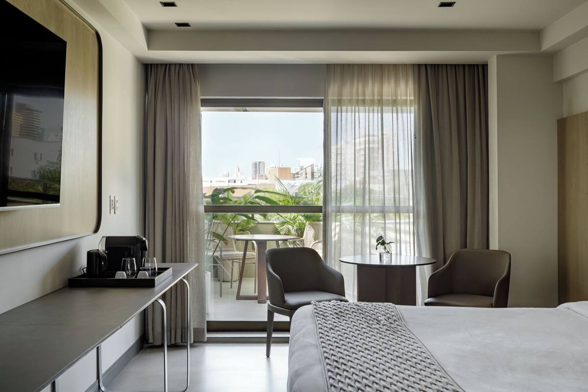 Una habitación de hotel moderna con una cama grande, un escritorio con cafetera, dos sillas, una mesita y un balcón con vistas a la ciudad.