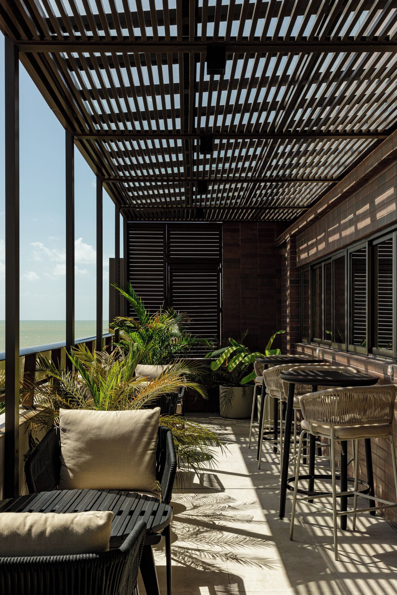 An outdoor seating area with a view of the ocean, featuring wooden pergola, wicker furniture, and potted plants.
