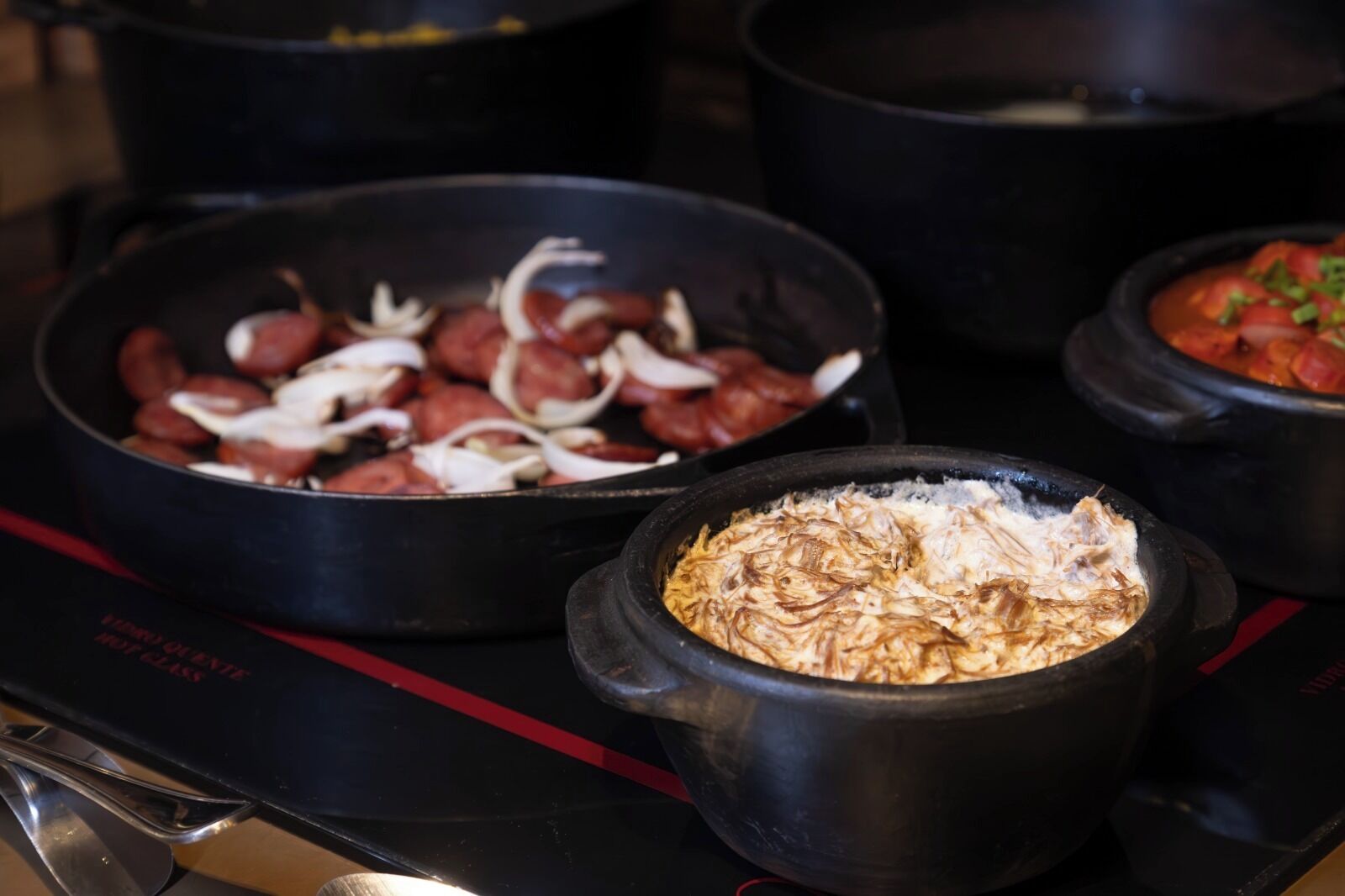 Eine Küche mit drei schwarzen Töpfen auf einem Herd, einer gefüllt mit Würstchen und Zwiebeln, ein anderer mit zerhacktem Fleisch und ein dritter mit einer Tomaten-basierten Speise.