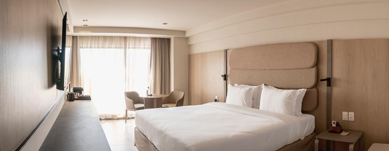 Habitación de hotel con una cama grande, un escritorio, dos sillas, un televisor montado en la pared y una ventana con cortinas.