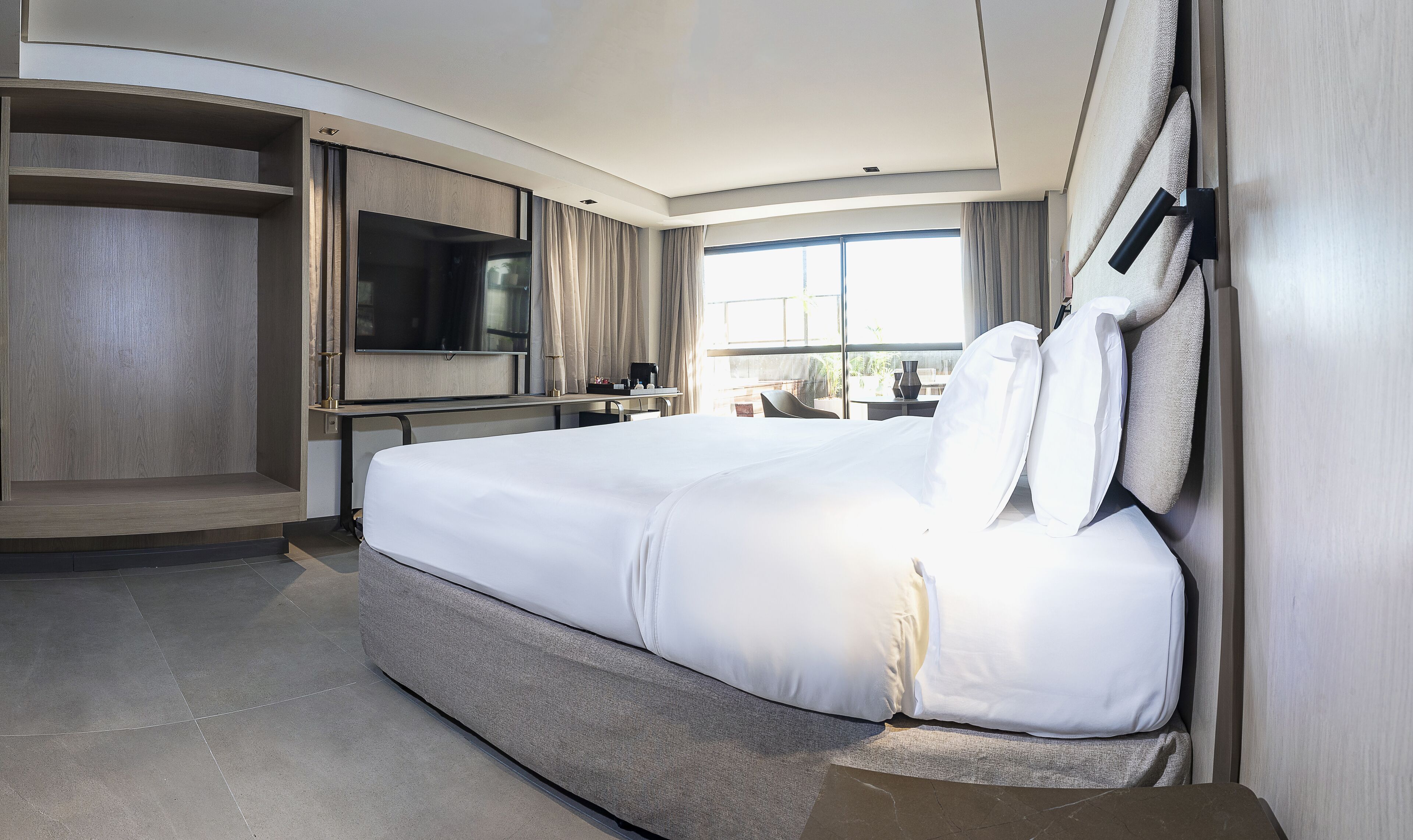 A modern hotel room with a large bed, a flat-screen TV mounted on the wall, a desk with a chair, and a window offering a view of the outdoors.
