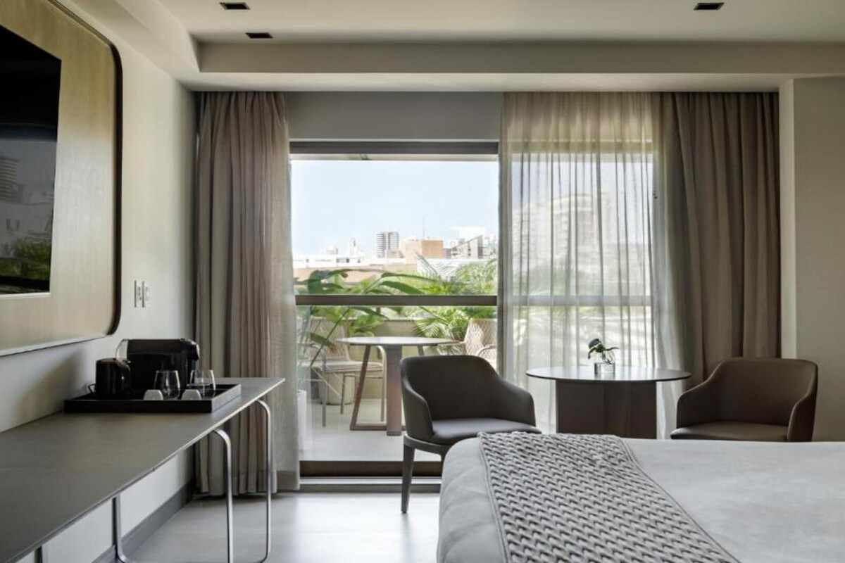 A hotel room with a bed, a desk, a chair, a small table, and a balcony with a view of buildings and greenery.