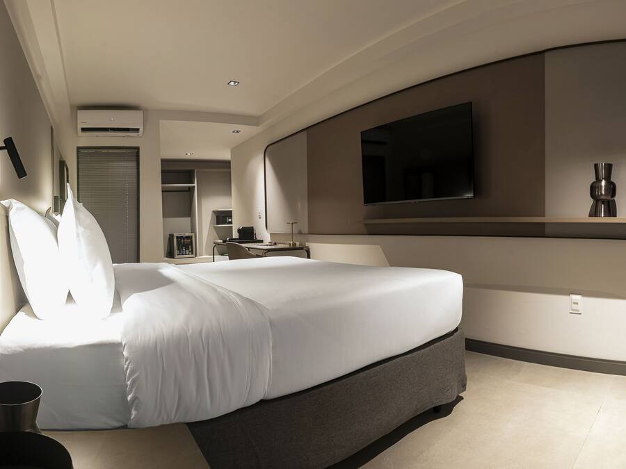 Quarto de hotel moderno com uma cama grande, televisão de tela plana na parede e uma pequena mesa com um vaso.