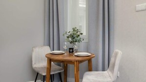 A dining area with a round wooden table, two white chairs, a vase with green foliage, and a window with curtains. - Cozy Studio in the Center | 1.3 km from the Main Square (Kraków)