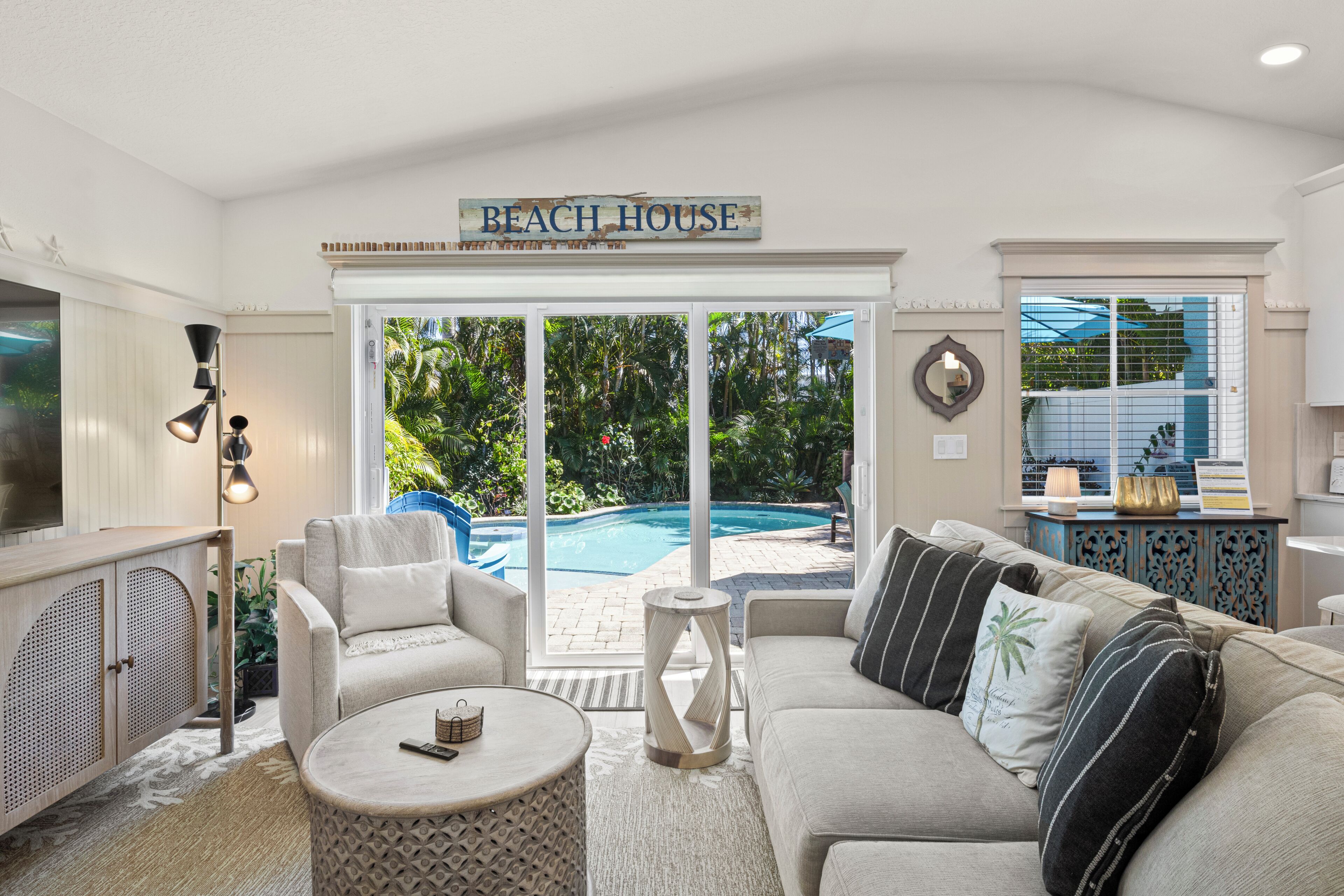 A living room with a pool view, a sign reading 'BEACH HOUSE,' and a cozy seating area.