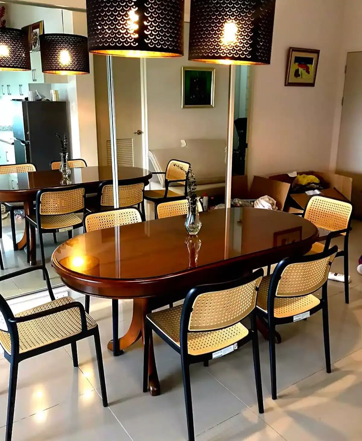A dining area with a large wooden table surrounded by wicker chairs, overhead pendant lights, and a vase on the table.