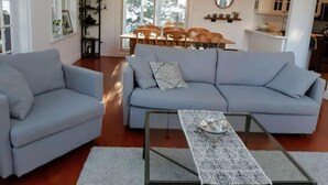 A modern living room with a grey sofa, a matching armchair, and a coffee table. There is a dining area with a wooden table and chairs, and a kitchen in the background.