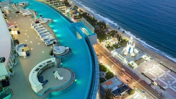 Un espace piscine aménagé sur le toit, avec des chaises longues et une fontaine circulaire, offrant une vue sur la plage et le paysage urbain.
