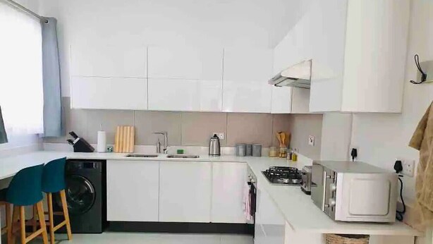 A modern kitchen with white cabinets, a sink, a stove, and a microwave. There are wooden stools and a basket on the counter. - Contemporary 2 bed in Suburbs - 2080 (Bulawayo)