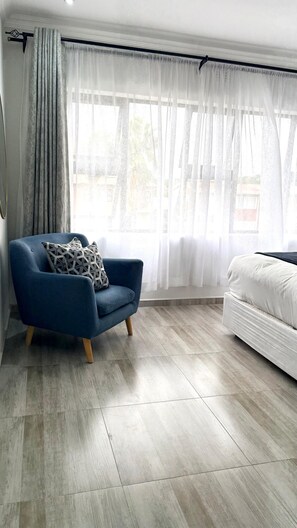 A room with a blue armchair, a white sofa, and a large window with sheer curtains. - Chic Modern House on Neasden - 2085 (Bulawayo)