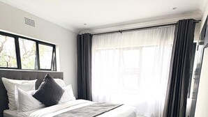 A modern bedroom with a large bed, a window with curtains, and a tiled floor. - Chic Modern House on Neasden - 2085 (Bulawayo)