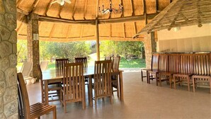 An outdoor covered dining area with wooden tables and chairs, a thatched roof, and a stone wall.