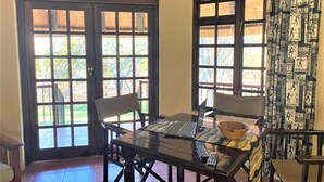 A room with a wooden table, two chairs, a laptop, and a bowl. There are two windows with curtains, and a door with glass panels.